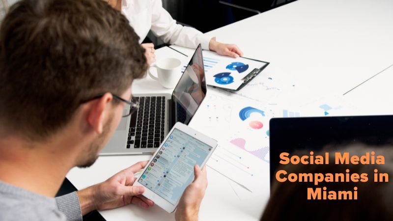 A group of professionals working at a table with laptops, tablets, and printed charts displaying data analytics. The text "Social Media Companies in Miami" is displayed in orange on the right side of the image.