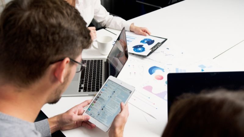 A group of professionals in a business meeting analyzing data on laptops, tablets, and printed charts. One person is holding a tablet displaying a social media platform, while another is using a laptop. A clipboard with data visualizations and printed graphs are spread across the table.