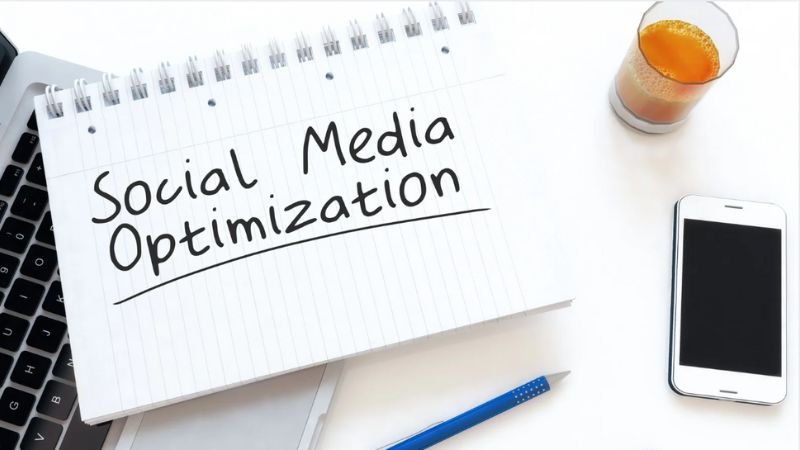 A notepad with "Social Media Optimization" written on it, placed on a laptop keyboard, alongside a smartphone, a blue pen, and a glass of orange juice on a white desk.